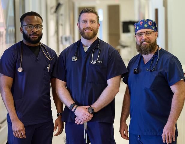 Three male nurses in navy blue scrubs stand in the hall