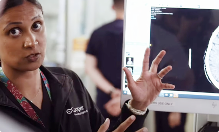 Clinician speaking and gesturing in front of a brain scan on a computer monitor.