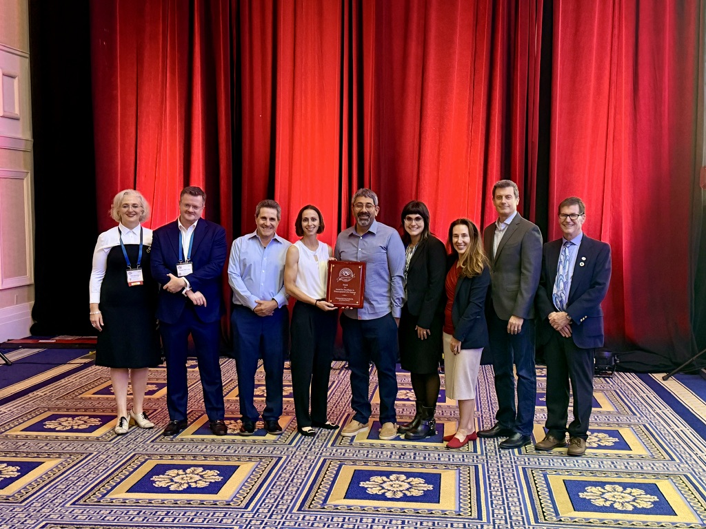 Representatives of the Cooper Critical Care Medicine team accepting Gold Center of Excellence by the Extracorporeal Life Support Organization (ELSO) for ECMO care award. Photo is a group of individuals against a red curtain backdrop with the center person holding an award plaque.  