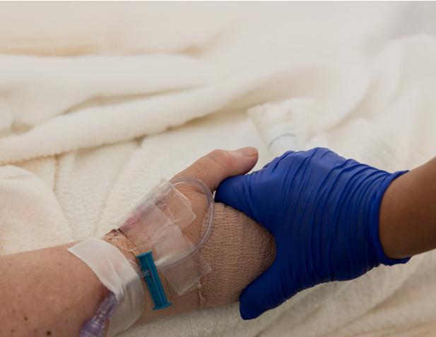 Close-up of a healthcare provider wearing a blue glove holding a patient’s bandaged hand with an IV line, offering comfort and support.
