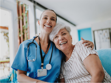 Smiling nurse with arm around an elderly woman, both seated and looking at the camera.