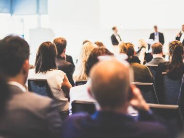 group of people in a conference room sit facing a panel on stage