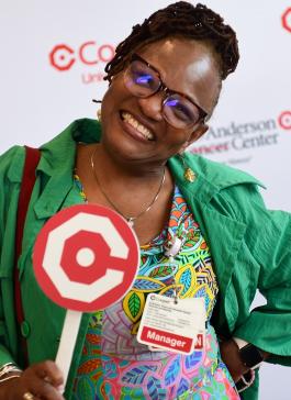 Black female employee in green jacket holds a Cooper logo on a stick and smiles at the camera