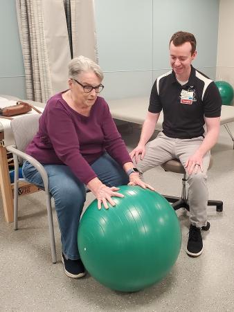 Female patient stretches arms to reach large green ball with male physical therapist watches